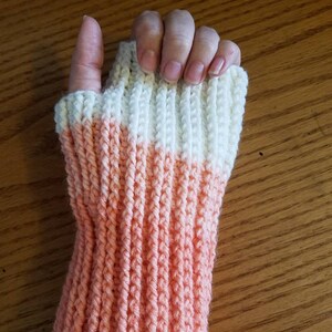 May include: A pair of knitted fingerless gloves with a peach and white color gradient. The gloves are made with a ribbed stitch.