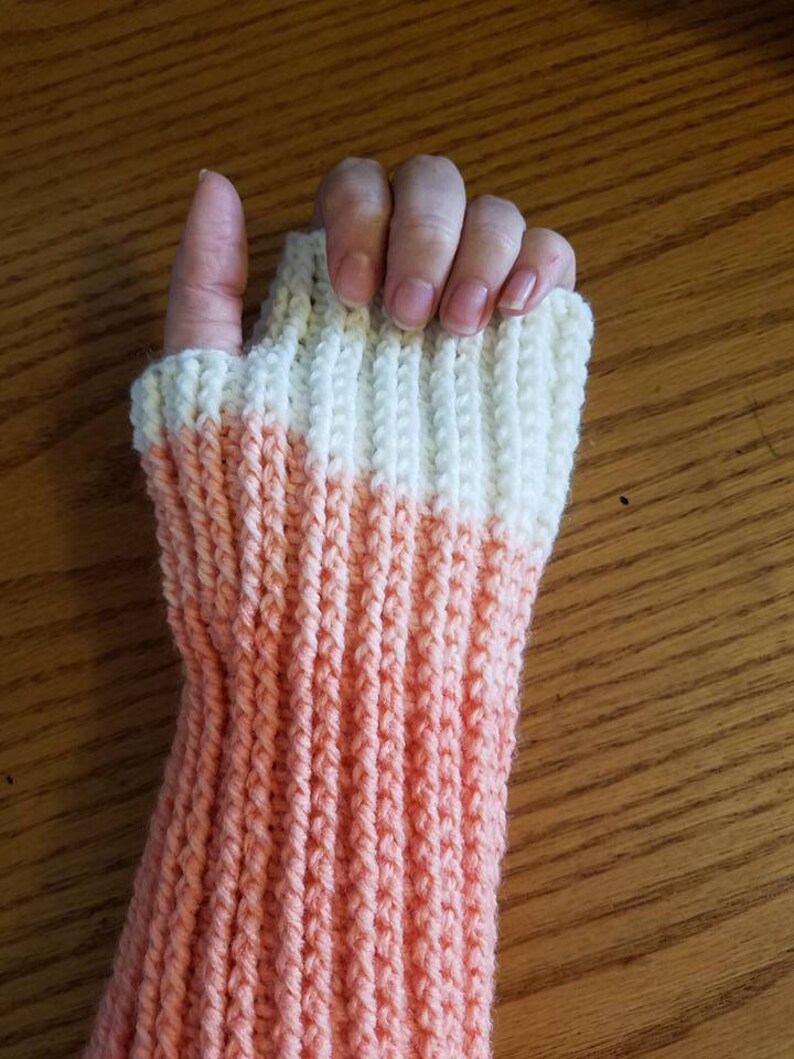 May include: A pair of knitted fingerless gloves with a peach and white color gradient. The gloves are made with a ribbed stitch.