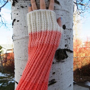 May include: A pair of knitted fingerless gloves with a white and coral color gradient. The gloves are made with a textured stitch and are shown against a white birch tree.