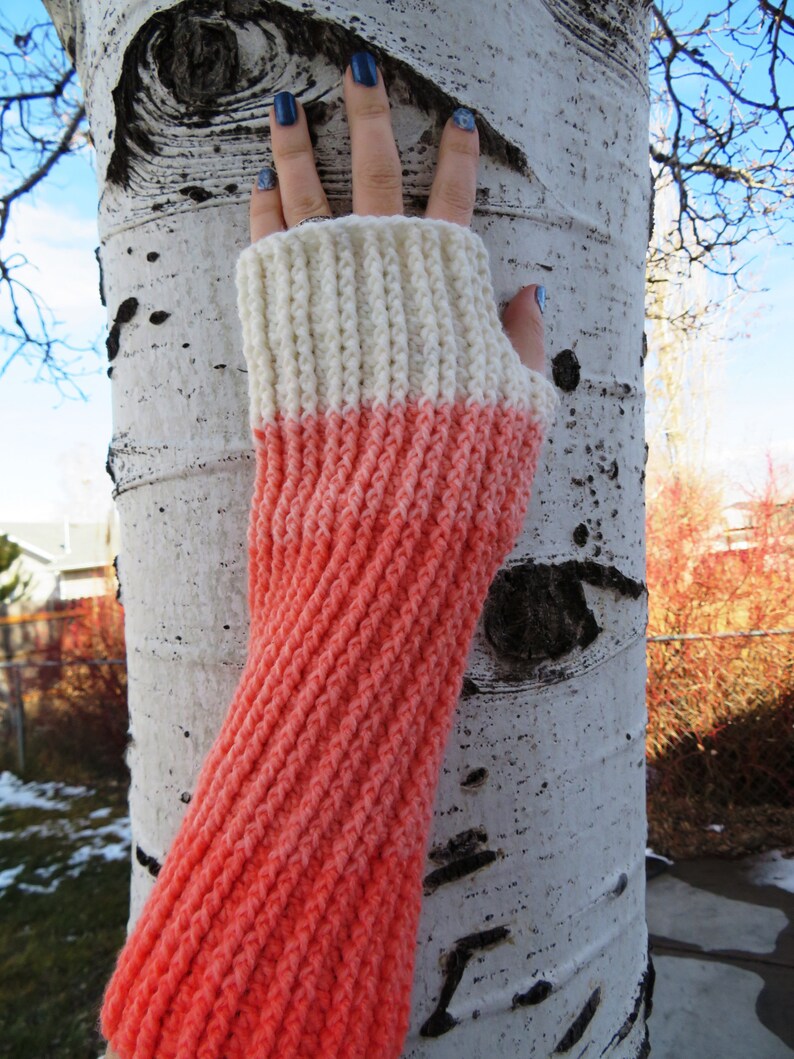 May include: A pair of knitted fingerless gloves with a white and coral color gradient. The gloves are made with a textured stitch and are shown against a white birch tree.