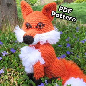 May include: A crocheted orange fox with white accents and large, expressive eyes sits amidst greenery and purple flowers. The fox has a black nose and paws, and a fluffy white chest. The text "PDF Pattern" is in the upper right corner.