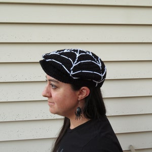 May include: A black and white crocheted hat with a spiderweb design. The hat is worn by a person with long dark hair and black spider earrings.