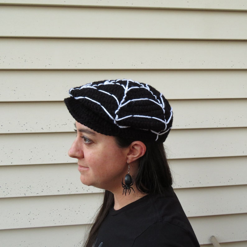 May include: A black and white crocheted hat with a spiderweb design. The hat is worn by a person with long dark hair and black spider earrings.