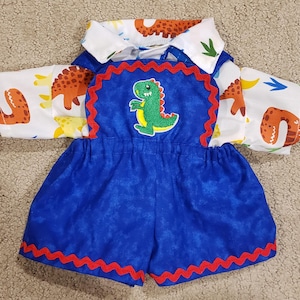 May include: A white short-sleeved shirt with a colorful dinosaur print worn under blue overalls. The overalls feature a green dinosaur patch on the chest, red rickrack trim along the edges of the bib and the hem of the shorts, and an elastic waist.