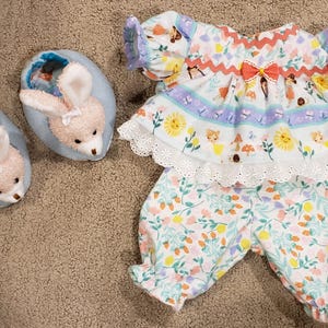 May include: A baby's floral print outfit with matching bunny slippers. The outfit includes a top with a lace trim and a bow, and matching bloomers. The slippers are light blue with bunny faces.