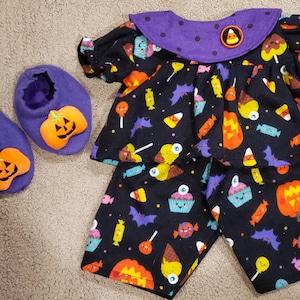 May include: A black doll outfit featuring a pattern of colorful Halloween candy, pumpkins, and bats, with a purple collar and a candy corn emblem. The set includes a short-sleeved top, matching pants, and two purple slippers with orange pumpkin appliques.