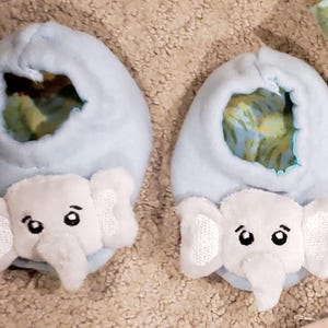 K&ouml;nnte beinhalten: Zwei hellblaue Babyschuhe mit wei&szlig;en Elefanten-Gesichtern. Die Schuhe haben eine weiche, pl&uuml;schige Textur und ein skurriles Design. Die Elefantengesichter haben gestickte Details, einschlie&szlig;lich Augen und Ohren. Die Innenseite der Schuhe hat einen farbenfrohen Stoff mit Muster.