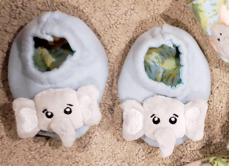 K&ouml;nnte beinhalten: Zwei hellblaue Babyschuhe mit wei&szlig;en Elefanten-Gesichtern. Die Schuhe haben eine weiche, pl&uuml;schige Textur und ein skurriles Design. Die Elefantengesichter haben gestickte Details, einschlie&szlig;lich Augen und Ohren. Die Innenseite der Schuhe hat einen farbenfrohen Stoff mit Muster.