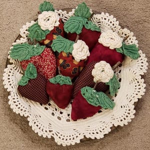 May include: A collection of handmade strawberry decorations in a wooden bowl atop a white lace doily. The strawberries are various shades of red and patterned fabric, with green crocheted leaves and white crocheted flowers.