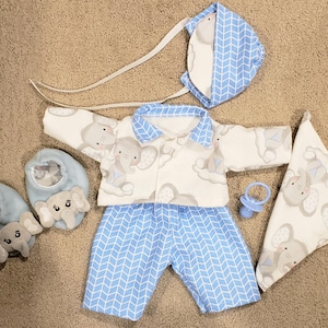 May include: A baby clothing set featuring a white and blue elephant print. The set includes a jacket, pants, bonnet, bib, and booties. A blue pacifier is also included. The booties have elephant faces.