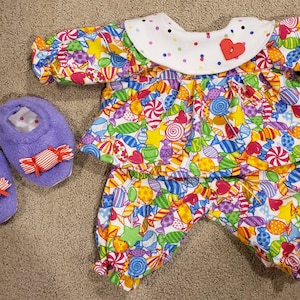 May include: A two-piece outfit consisting of a long-sleeved top and pants featuring a colorful candy print on a white background. The top has a white collar with multi-colored dots and a red heart button. Two purple plush slippers with candy accents.