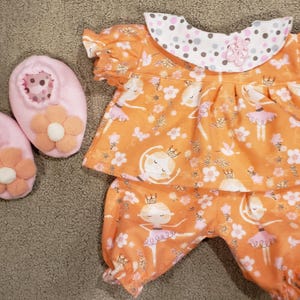May include: An orange doll outfit with a ballerina print, including a top, pants, and two pink shoes. The top has a white collar with pink and gray polka dots. The shoes have orange flower accents.