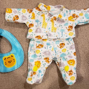 May include: A baby's pajama set and bib. The white pajamas have a colorful jungle animal print, including tigers and giraffes. The light blue bib has a tiger face applique.