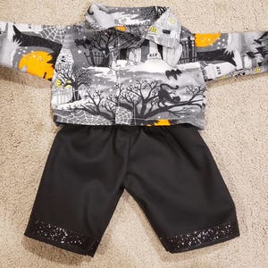 May include: A long-sleeved button-down shirt featuring a gray, black, and orange Halloween-themed print with tombstones, trees, and cats, paired with solid black shorts that have a black glittery band along the bottom hem of each leg.