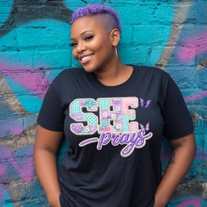 Könnte beinhalten: Schwarzes T-Shirt mit dem Text "SHE prays" in einem Patchwork-Design mit floralen und Schmetterlingsakzenten. Das T-Shirt wird vor einer farbenfrohen Graffiti-Wand getragen.