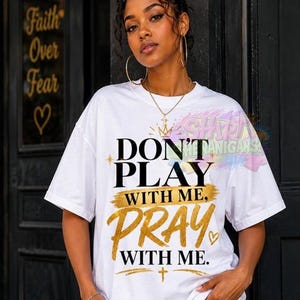 Może przedstawiać: Biały t-shirt z krótkim rękawem z czarno-złotym napisem "DON'T PLAY WITH ME, PRAY WITH ME." Wzór zawiera złote akcenty w formie pociągnięć pędzla, małą złotą koronę, serce oraz symbol krzyża nadrukowany na przodzie ubrania.