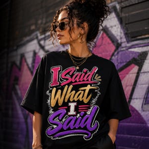 May include: A black short-sleeved t-shirt featuring the text I Said What I Said in a stylised graphic design. The word I is in pink, Said is in pink, What is in gold, I is in white, and Said is in purple with a brushstroke effect.