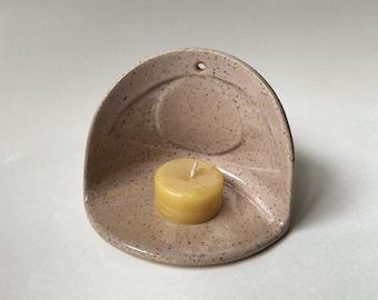 May include: A speckled light brown ceramic wall hanging with a small round indentation for a candle. A yellow candle is placed in the indentation.