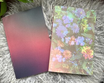 May include: Two notebooks, one with a gradient of dark purple to red and the other with a floral pattern in shades of blue, green, and pink.