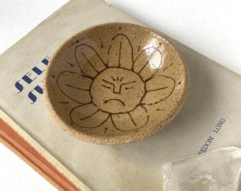 May include: A small, brown ceramic dish with a speckled glaze. The dish is decorated with a brown line drawing of a flower with a face. The flower has a frowning expression.
