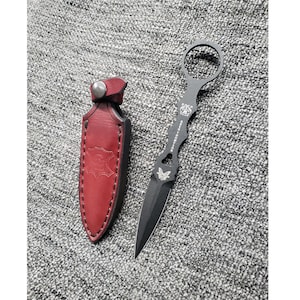 May include: A black tactical knife with a butterfly design and a ring handle. The knife is in a red leather sheath.