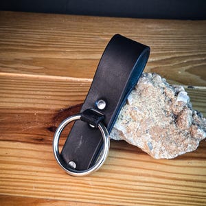 May include: A black leather keychain with a large silver ring. The leather strap is riveted and attached to the ring. The keychain is resting on a wooden surface next to a rock.