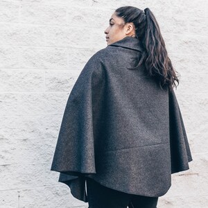 Puede incluir: Una capa de lana gris con cuello alto. La capa la lleva puesta una persona con el pelo largo y oscuro recogido en una coleta.