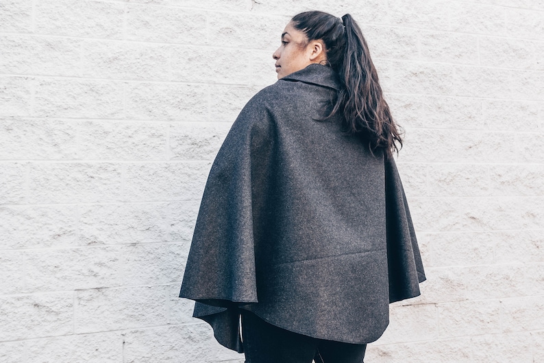 Puede incluir: Una capa de lana gris con cuello alto. La capa la lleva puesta una persona con el pelo largo y oscuro recogido en una coleta.