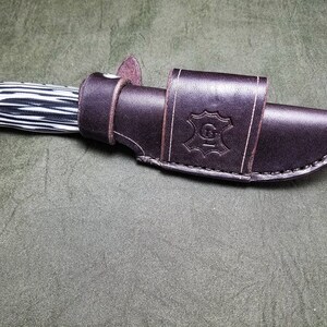 May include: A brown leather knife sheath with a black and white patterned knife inside. The sheath has a stitched design and a leather logo with the letter "G" in the center.