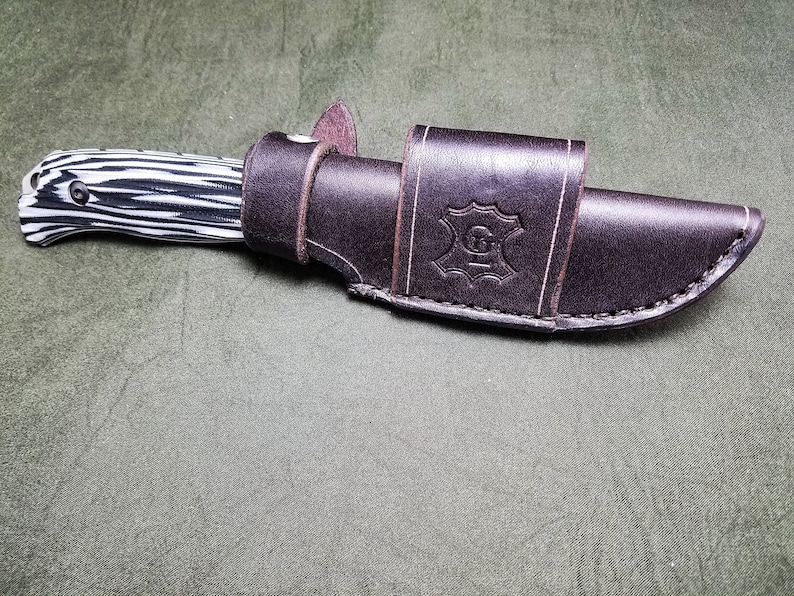 May include: A brown leather knife sheath with a black and white patterned knife inside. The sheath has a stitched design and a leather logo with the letter "G" in the center.