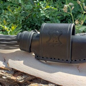 May include: A black leather knife sheath with a stitched design and a small leather tag with the text "100% Leather" embossed on it. The sheath is holding a black and white patterned knife.