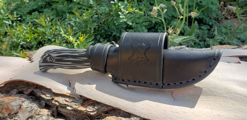 May include: A black leather knife sheath with a stitched design and a small leather tag with the text "100% Leather" embossed on it. The sheath is holding a black and white patterned knife.