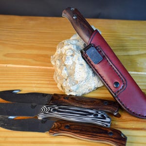Puede incluir: Un surtido de cuchillos con mangos de madera y hojas negras. Un cuchillo está en una funda de cuero roja. Los cuchillos están dispuestos sobre una superficie de madera, con una piedra en el fondo.