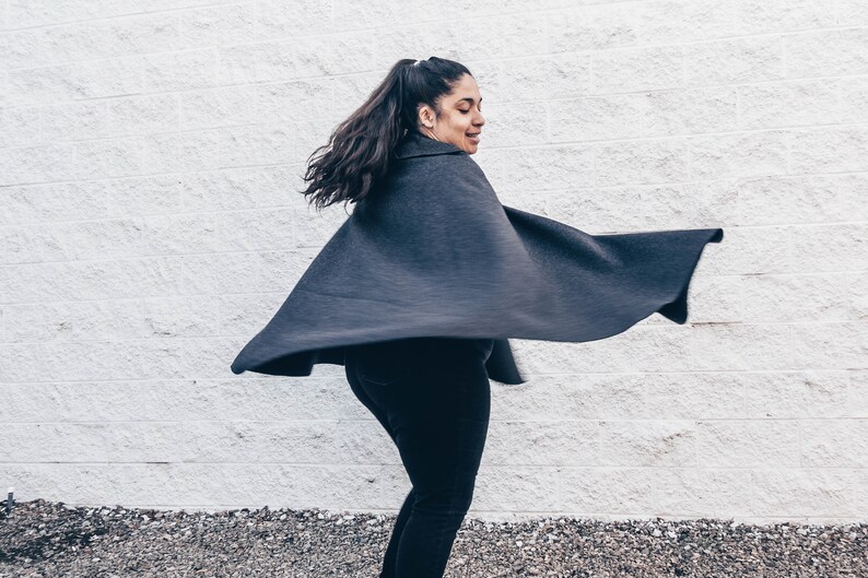 Puede incluir: Una mujer con una capa negra y una larga cola de caballo oscura. La capa ondea con el viento y la mujer est&aacute; de pie frente a una pared de ladrillo blanco.