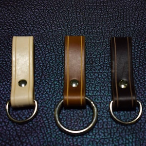 May include: Three leather key fobs with metal rings. The fobs are in different colours: beige, brown, and dark brown.