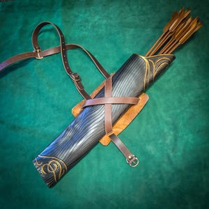 May include: A black quiver with a textured, wavy surface and gold-painted decorative accents, featuring a brown leather harness system with adjustable buckles and straps. Several wooden arrows with fletching are stored inside the quiver.