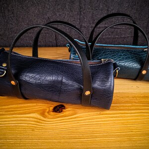 Puede incluir: Dos peque&ntilde;as bolsas de cuero cil&iacute;ndricas con correas negras y detalles dorados. La bolsa de la izquierda es azul oscuro y la bolsa de la derecha es azul verdoso.