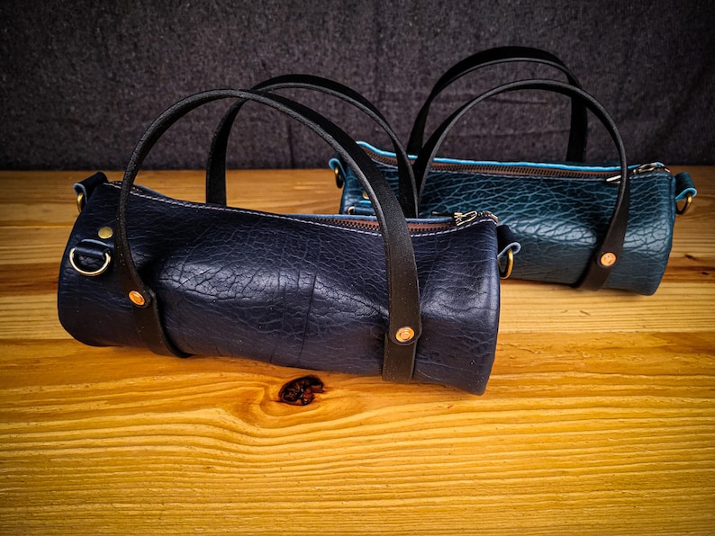 Puede incluir: Dos peque&ntilde;as bolsas de cuero cil&iacute;ndricas con correas negras y detalles dorados. La bolsa de la izquierda es azul oscuro y la bolsa de la derecha es azul verdoso.