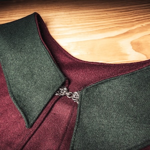 Puede incluir: Primer plano de una prenda de lana color burdeos y verde con un cierre met&aacute;lico decorativo. La prenda tiene un cuello con un borde puntiagudo.