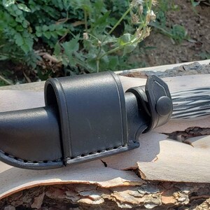 May include: A black leather knife sheath with a snap closure. The sheath is designed to fit a knife with a black and white patterned handle.