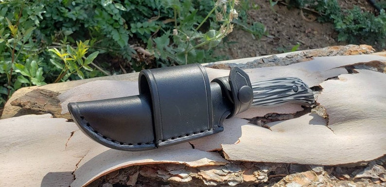 May include: A black leather knife sheath with a snap closure. The sheath is designed to fit a knife with a black and white patterned handle.