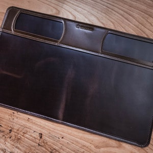 May include: A brown leather desk mat with two rectangular cutouts for holding business cards or other small items. The mat has a stitched border and the text "Saddleback" is embossed in the upper right corner.