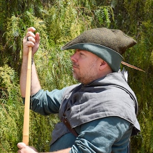 以下が含まれることがあります： 緑と茶色の、中世風の帽子とチュニックを着た人が、木の弓を持っています。帽子は濃い緑色のつばと羽が付いています。背景は緑豊かな緑の葉です。