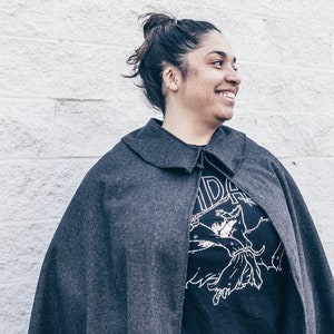 Puede incluir: Una mujer con una capa gris sobre una camiseta negra. La camiseta tiene un estampado gr&aacute;fico de una criatura con los brazos extendidos. La mujer sonr&iacute;e y mira hacia la derecha.