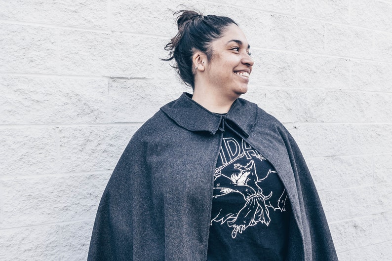 Puede incluir: Una mujer con una capa gris sobre una camiseta negra. La camiseta tiene un estampado gr&aacute;fico de una criatura con los brazos extendidos. La mujer sonr&iacute;e y mira hacia la derecha.