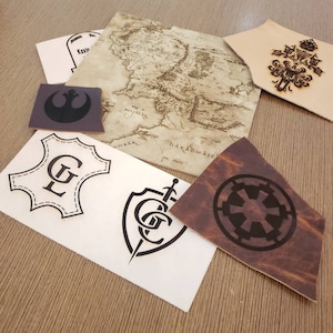 May include: A collection of leather patches with various designs, including a map, a shield with a sword, a star wars symbol, and a monogram. The patches are on a wooden surface.