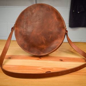 May include: A round, brown leather shoulder bag with a long, adjustable strap. The bag has a distressed finish and a simple, classic design. The strap is attached with brass-colored hardware. The bag is sitting on a wooden surface.