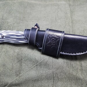 May include: A black leather knife sheath with a white stitching detail and the letter "G" embossed on the front. The sheath is holding a black and white patterned knife.