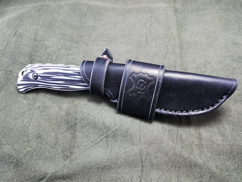 May include: A black leather knife sheath with a white stitching detail and the letter "G" embossed on the front. The sheath is holding a black and white patterned knife.