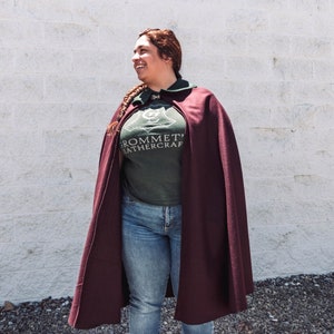 Puede incluir: Una mujer con una capa granate con una camiseta verde que dice "Grommet Leathercraft" y un emblema dorado. Est&aacute; de pie frente a una pared de ladrillo blanco.